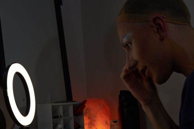 A drag queen sits in front of a vanity mirror and puts makeup on her face.