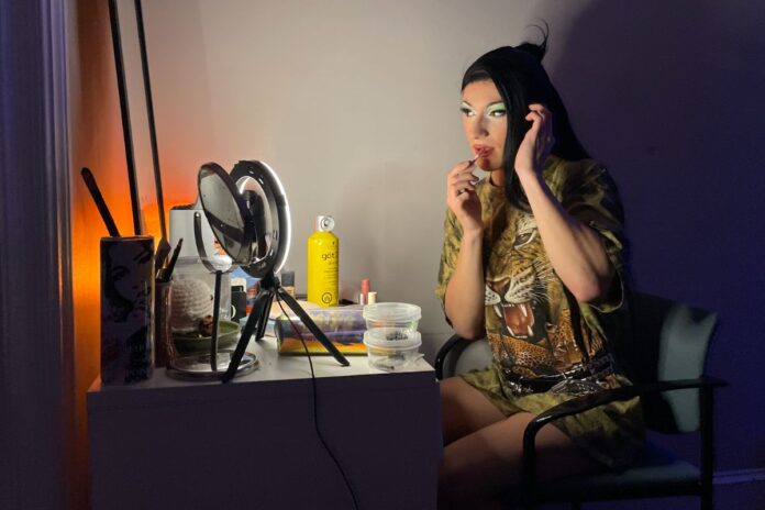 A drag queen puts lip gloss on her lips in front of her vanity mirror.