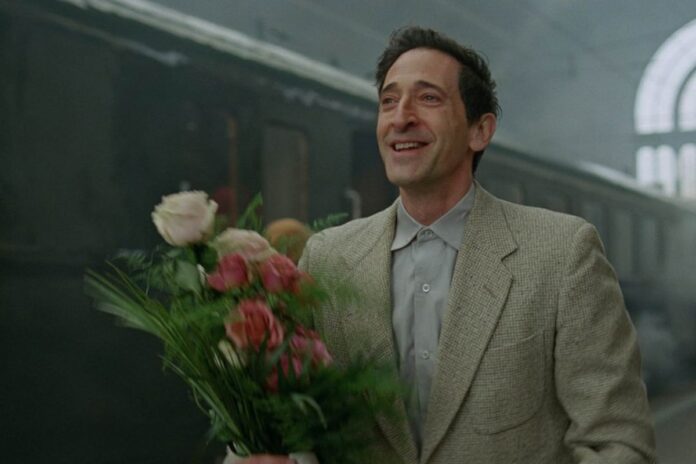 Adrien Brody as László Tóth in 'The Brutalist' wearing a tan suit and holding flowers beside a train.