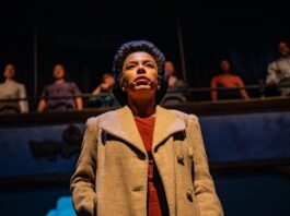 Woman stands on stage in brown trenchcoat