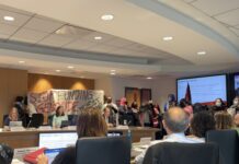 A group of students hold a banner reading, "stop funding genocide" in a room of people.