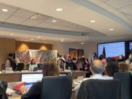 A group of students hold a banner reading, "stop funding genocide" in a room of people.