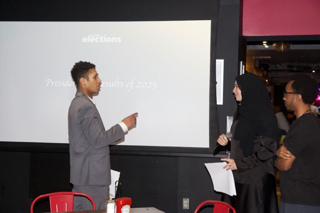 A man stands and speaks while facing a woman and another man.