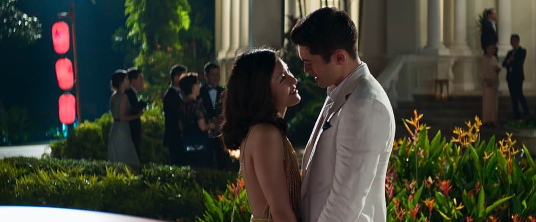 A woman and a man stand holding hands, staring at each other, in front of a garden and a group of people in the background.