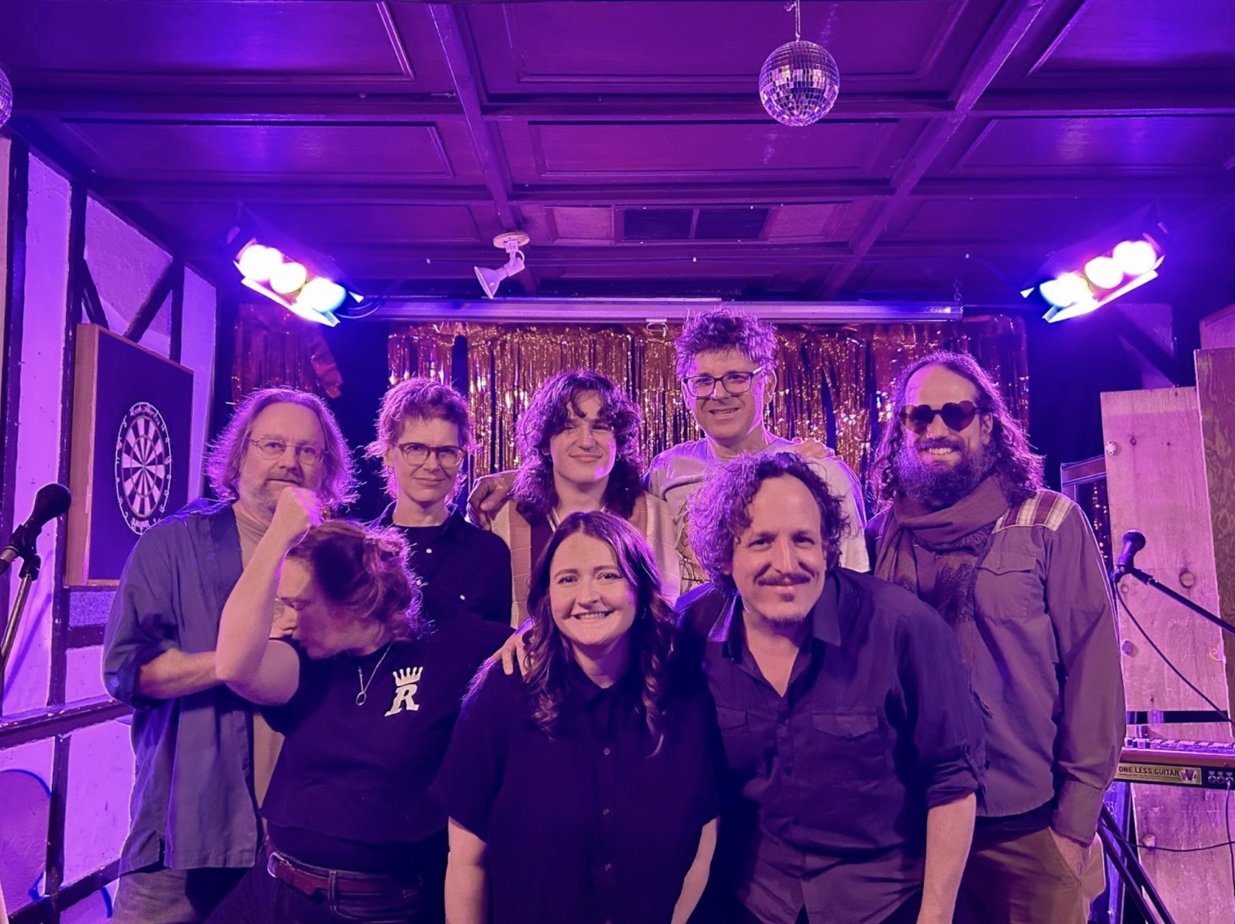 A group of people stand together, smiling, in a room with purple lighting. 