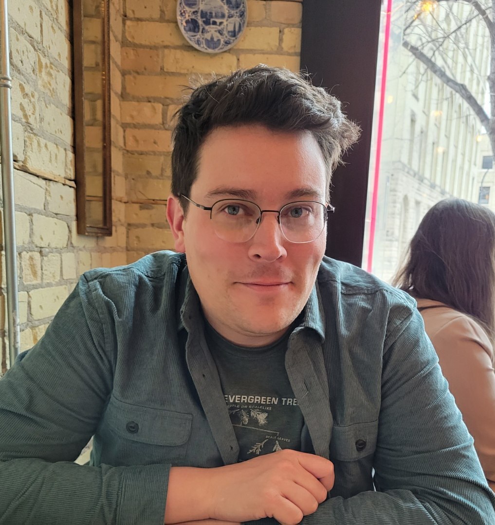 A person with dark hair and wearing glasses sits in front of a brick wall and window.