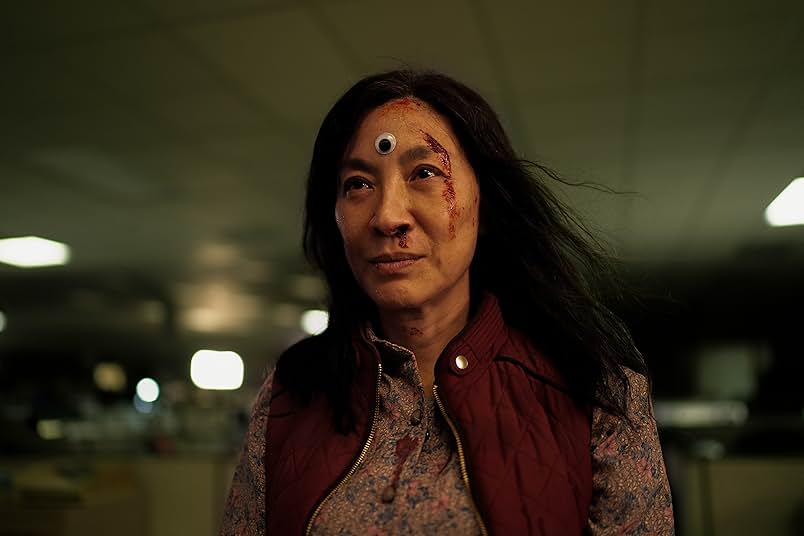 A woman with a plastic googly eye on her forehead stands and looks into the distance.