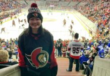 A person wearing an Ottawa Senators jersey and hat stands in a crowded arena while the Ottawa Senators warm up on the rink.