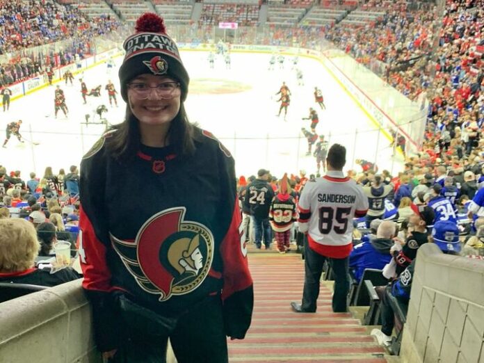 Sens-playoffs-feature A person wearing an Ottawa Senators jersey and hat stands in a crowded arena while the Ottawa Senators warm up on the rink.