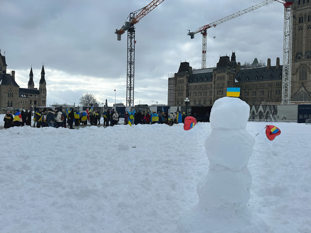 A small snowman with a Ukrainian flag sits by Parliament Hill, with protesters in the background supporting Ukraine.
