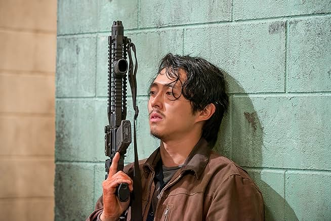 A person with black, sweaty hair holds a gun and stands against a blue wall.