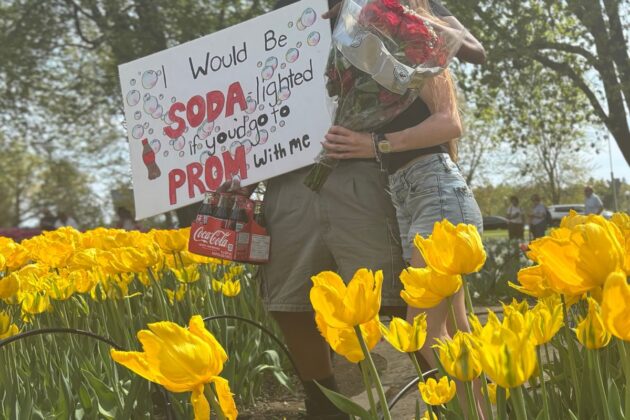 A couple stand in a field of tulips, holding a prom proposal sign and a bouquet of flowers.