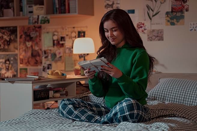A young girl wearing blue PJ bottoms and a green sweater sits on a bed holding a stack of letters.