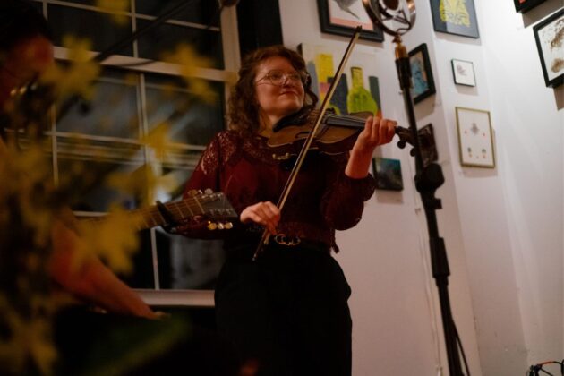 A person plays the fiddle in a café with framed photos on the wall in the background.