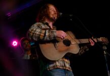 A person wearing a plaid shirt and holding a guitar behind a mic stand performs in a darkly-lit venue.