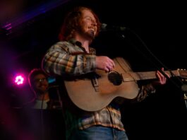 A person wearing a plaid shirt and holding a guitar behind a mic stand performs in a darkly-lit venue.