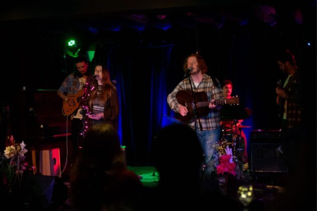 In a darkly-lit venue, a four-piece band perform with instruments on stage.