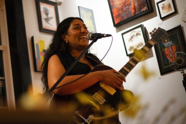 A person smiles into a mic stand while holding a guitar and performing in a cafe with framed photos on the wall in the background.