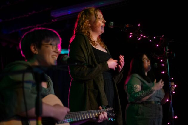 Three people behind mic stands sing and perform in a darkly-lit venue.