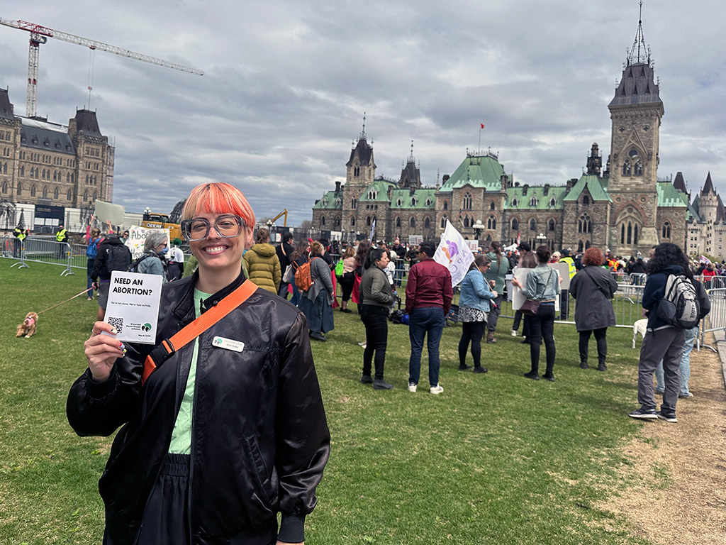 A person with coloured hair stands in front of a crowd of protesters, holding up a pamphlet.