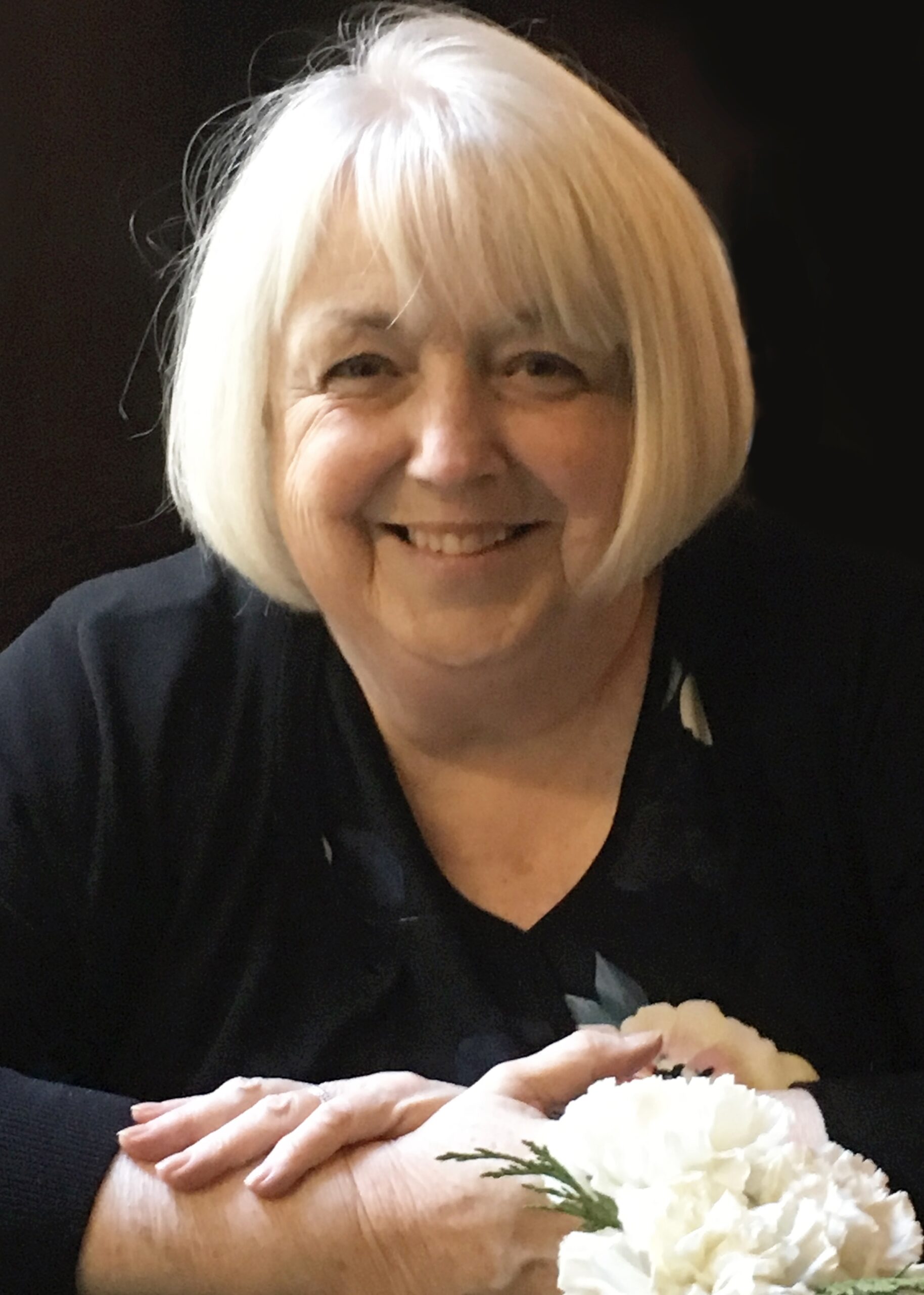 a woman with white hair, folded hands around white flowers, smiles at the camera