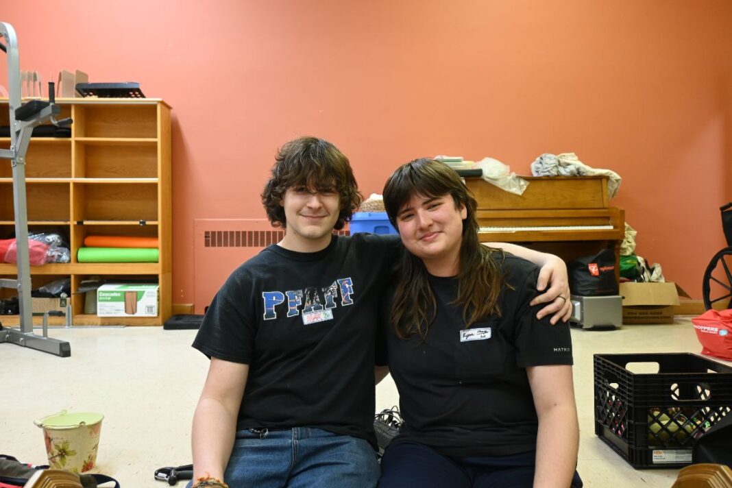 Two people wearing black sit in a room, with their arms around each other.