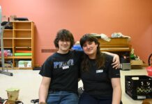 Two people wearing black sit in a room, with their arms around each other.