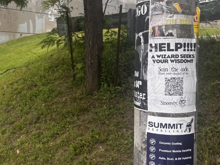 A lamp post stands in front of a grassy hill, it has posters taped to it. 