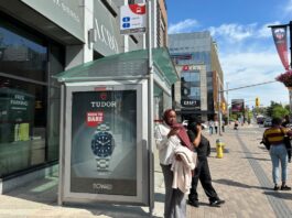 A person wearing a white shirt stands outside a bus stop, on their phone, seemingly waiting for a bus.