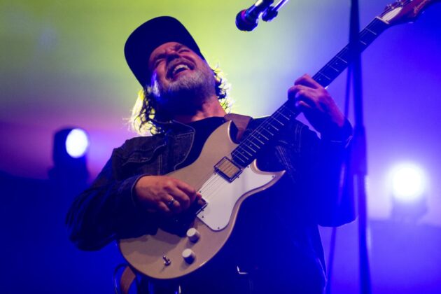 A man wearing a hat and a black shirt sings passionately while playing guitar.