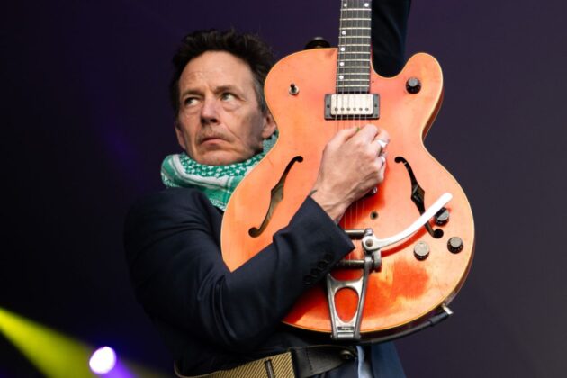 A person wearing black and a green scarf holds up a guitar with a serious-looking face.