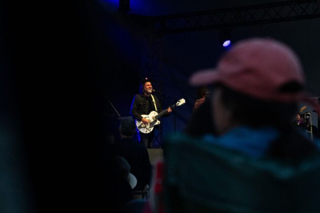 The crowd looks upon a guitar player in the distance on a black stage.