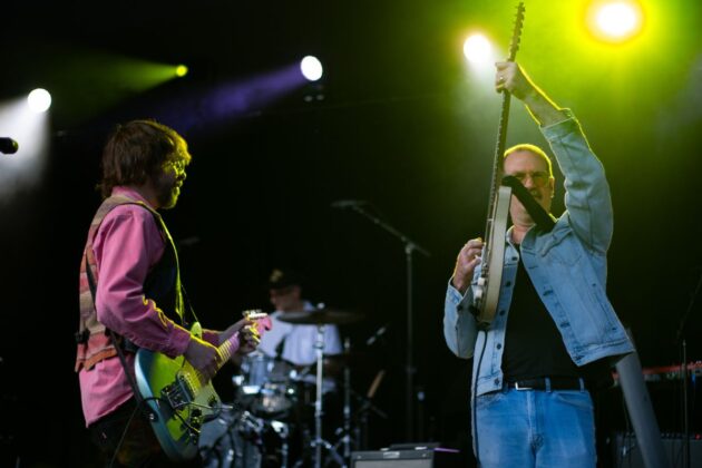 A band performs on stage, two men are in the front with guitars under a green spotlight.