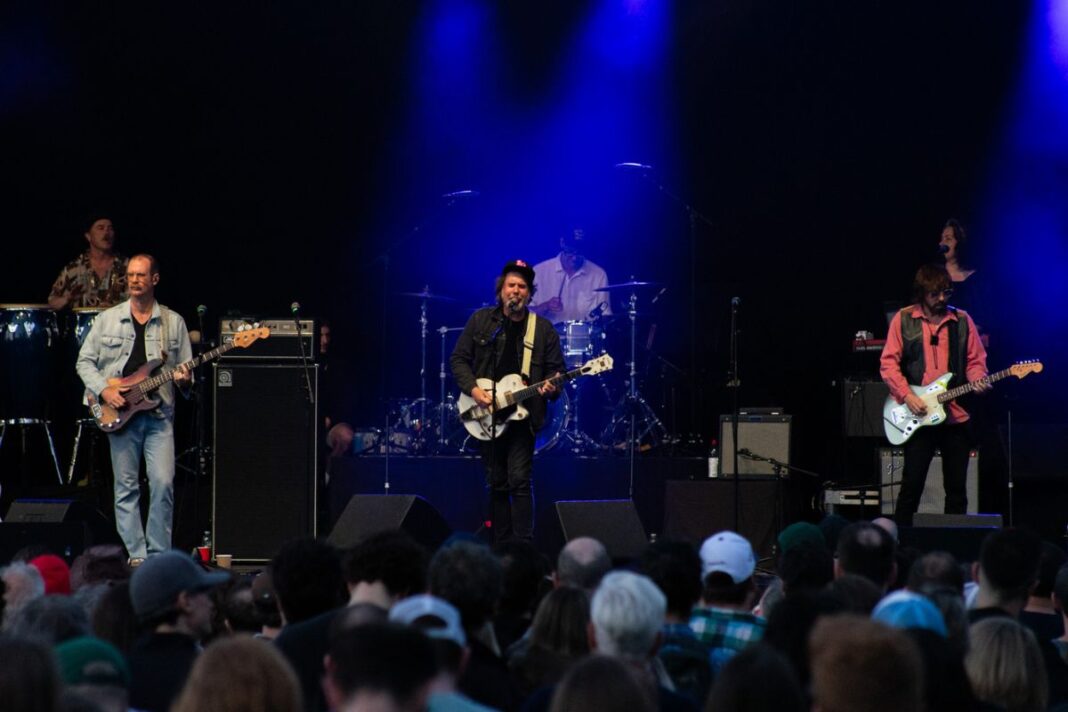 A band with guitars, drums and other instruments stand on a stage backlit with blue.