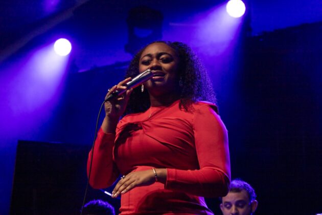 A singer in a red dress holds up a mic and sings on stage in purple lighting.