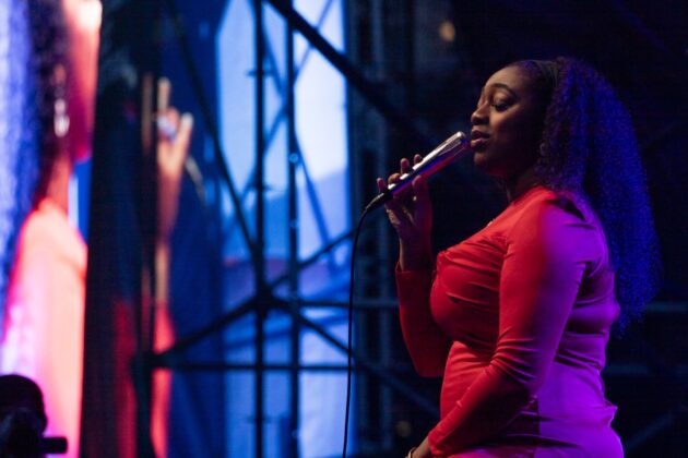 A singer in a red dress sings on stage, next to a screen projecting her performance at night.