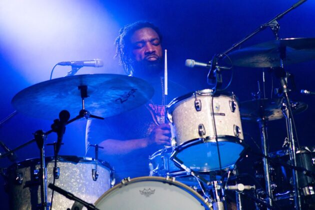 A drummer plays drums behind a drum kit, cast in blue lighting.