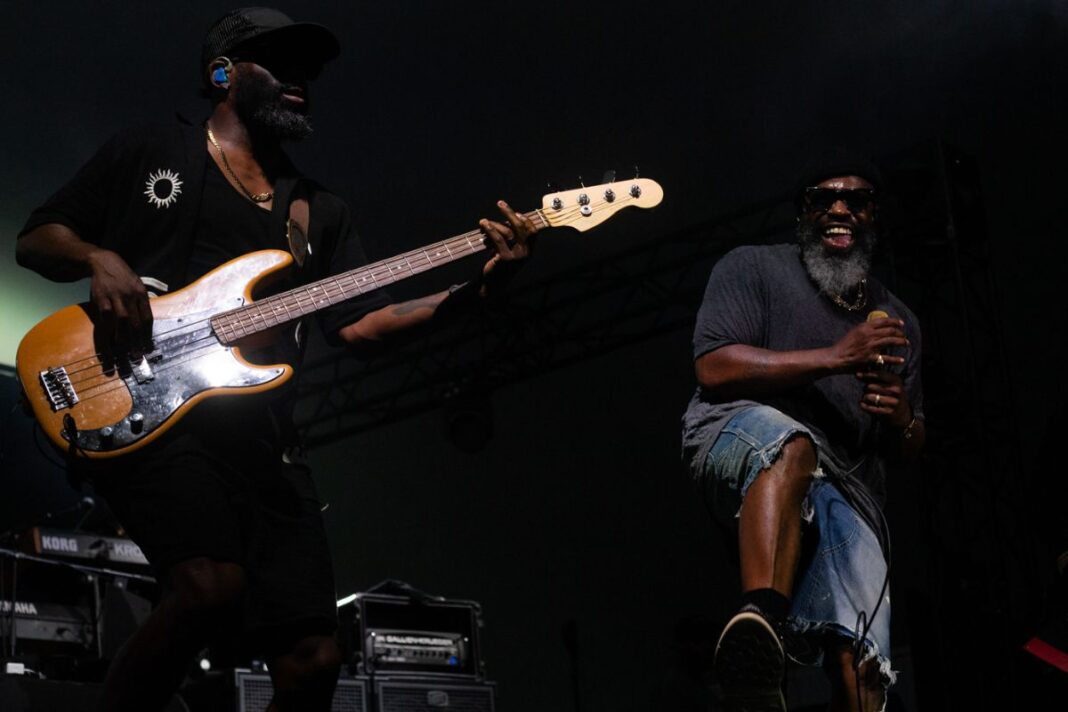 Two men stand on stage, one has a bass guitar and the other grins.