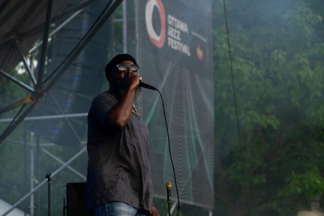 A person sings on stage as the sun sets, in front of an Ottawa Jazz Festival banner.
