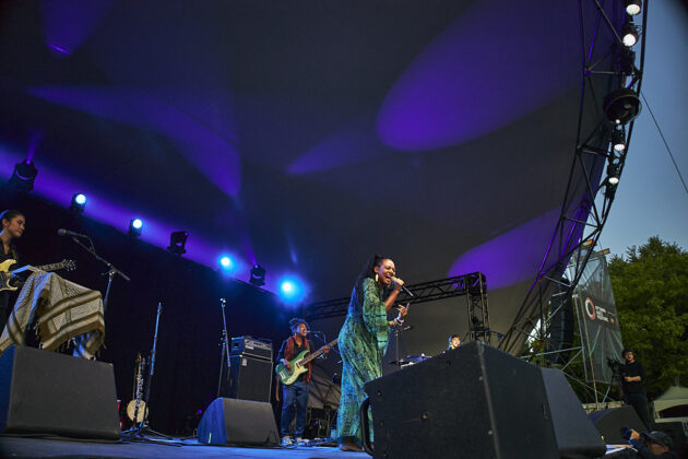 A woman sings on stage with purple and blue backlighting as the sun begins to set.