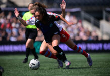 Two soccer players, one behind and one in front, fight for the ball on a field.