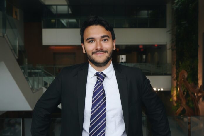 Artur Award A person wearing a suit and tie stands and smiles, backdropped by a large space.