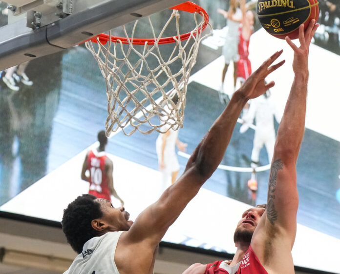 Player (Zane Waterman) lifts basketball in the air to make a layup with another player reaching across to try and block the shot.