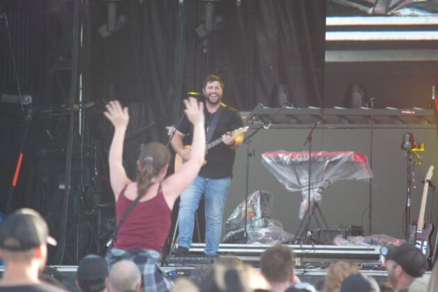 A man in blue jeans and a black shirt plays guitar and smiles at a crowd, a person in the crowd is sitting on someone's shoulders