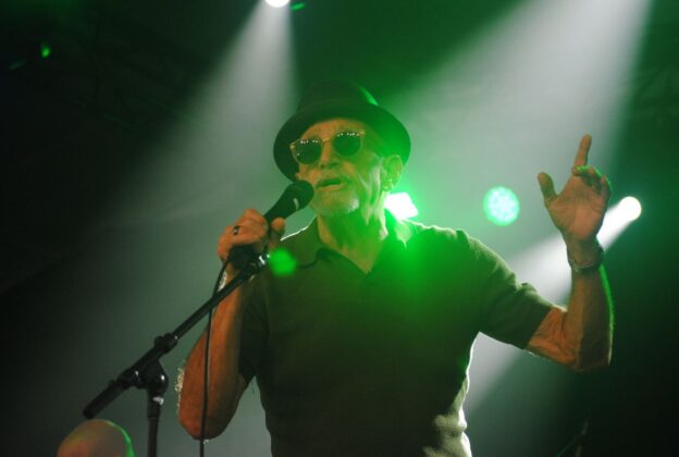 A man in a black hat and sunglasses sings on a stage under green lighting