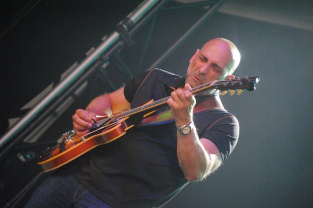 A guitarist in a black outfit plays on a stage.