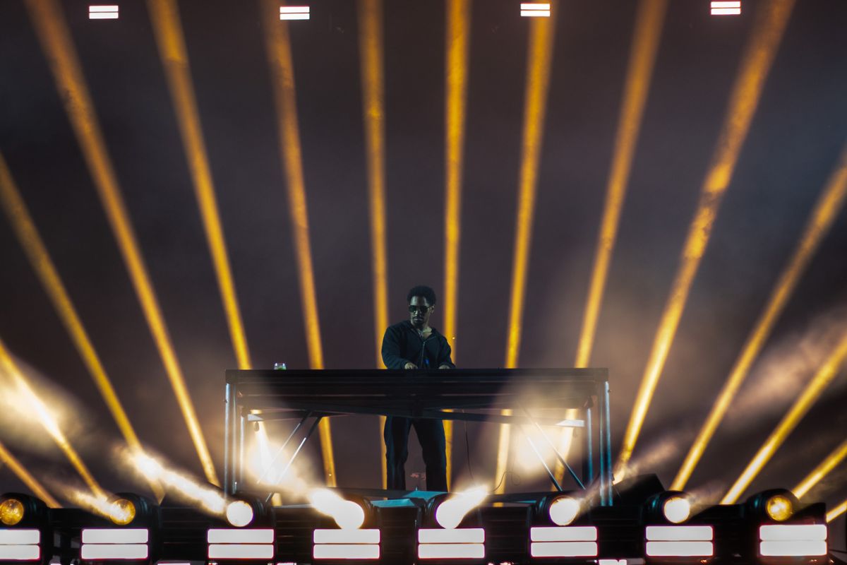 A DJ performs on a stage with a simple, black backdrop and yellow spotlights.