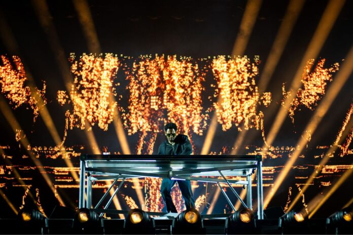 A DJ plays on stage in with a backdrop of golden spotlights and sparks.