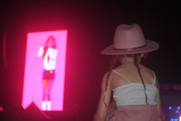 A child in a pink cowgirl hat sits on the shoulders of her parents at a concert.