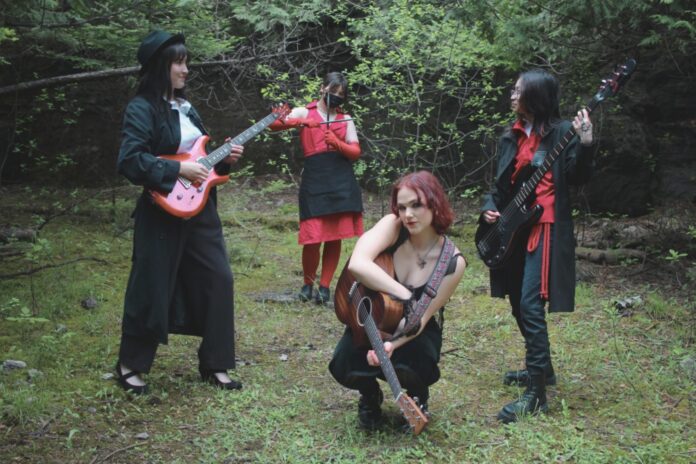 Four people in red and black extravagant clothing stand with instruments in a forest clearing.